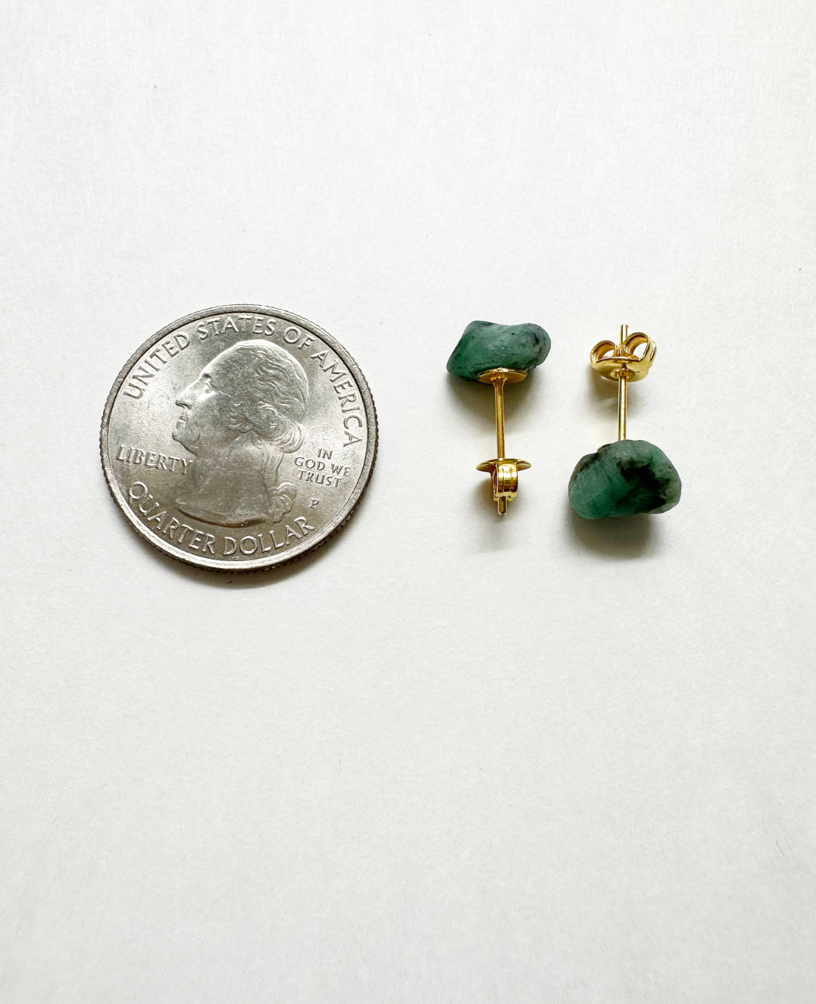 Raw emerald stud earrings next to a quarter for size comparison.