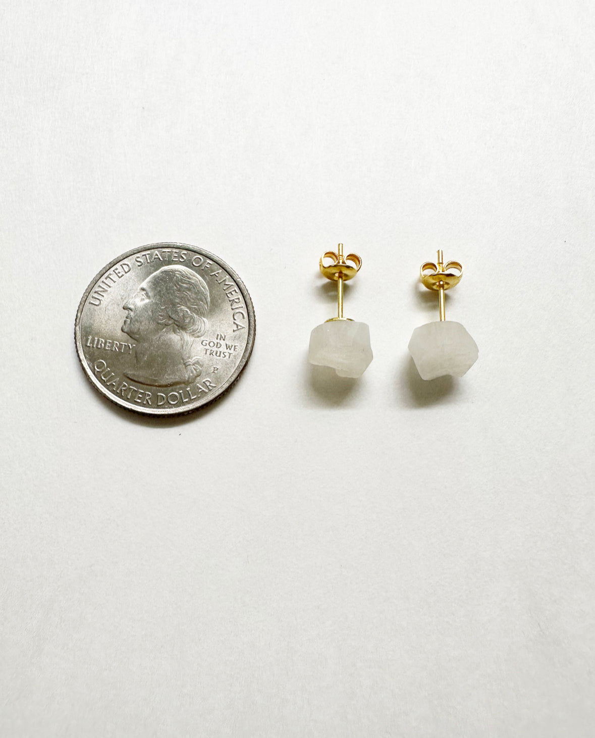 Raw rainbow moonstone stud earrings next to a quarter for size comparison.